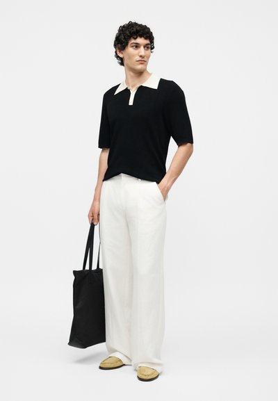 Άνδρας με σγουρά μαλλιά που φοράει μαύρο πόλο με κοντά μανίκια, λευκό φαρδύ παντελόνι, μπεζ παπούτσια, κρατώντας μια μαύρη τσάντα tote, στέκεται μπροστά σε απλό φόντο.