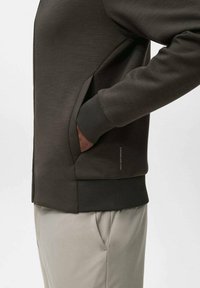 Chaqueta soft-shell de color marrón oscuro con cremallera, bolsillos laterales, puños acanalados y un diseño ajustado. Tela texturizada con un acabado mate.