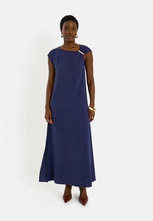 Femme portant une robe maxi bleu marine sans manches avec une fente latérale, des chaussures pointues marron, des boucles d’oreilles créoles dorées et des bracelets.