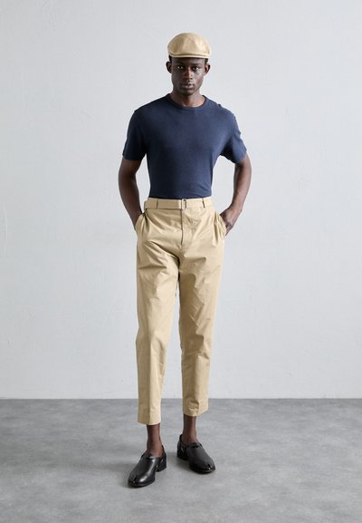 Chemise bleu marine à manches courtes, pantalon taille haute beige et chaussures noires à enfiler. La casquette est d'un beige clair, complétant le look structuré.