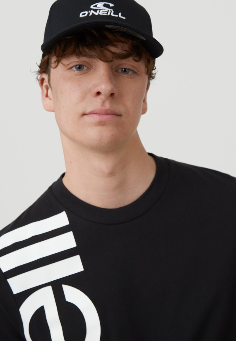 Sort t-shirt med stort hvidt grafisk logo og striber på venstre skulder, parret med en sort kasket med hvidt mærkelogo.