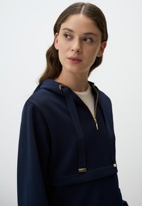 Hoodie blu scuro con zip, caratterizzato da hardware dorato, un tessuto testurizzato e cordini regolabili sul cappuccio.