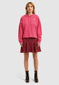 Roze hoodie met een cropped ontwerp, gecombineerd met een uitlopende bloemenrok in tinten rood en roze, en glanzende zwarte enkellaarsjes.