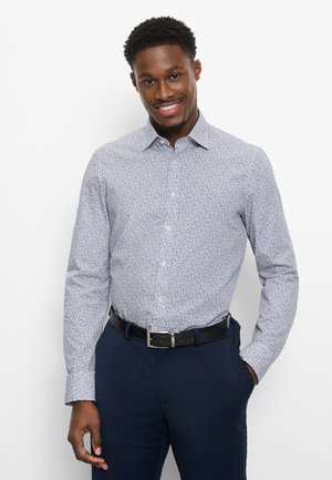 Mann lächelt, trägt ein weiß gemustertes langärmeliges Hemd, in dunkelblaue Hose mit schwarzem Gürtel gesteckt, eine Hand in der Tasche.