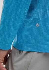 Sininen pitkähiha paita, jossa pieniä valkoisia pilkkuja ja hopeinen logo vasemman alareunan kohdalla. Harmaat shortsit näkyvissä alhaalla.