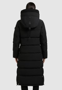 Jachetă puffer neagră, cu un glugă mare, detașabilă, detalii ribbate și accente cu fermoar. Are o siluetă lungă, ușor cambrată, cu buzunare.