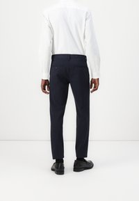 Man in marineblauwe streeppantalons, zwarte loafers en een witte overhemd met lange mouwen en knopen, staand tegen een effen witte achtergrond.