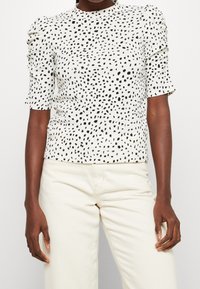 Top bianco con motivo a macchie nere, maniche corte e dettaglio arricciato sulle spalle. Abbinato a pantaloni di colore chiaro.