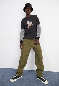 Camiseta gráfica negra de manga larga con mangas a rayas, pantalones cargo verdes y sombrero tipo cubo negro. Zapatillas negras y blancas con un diseño clásico.