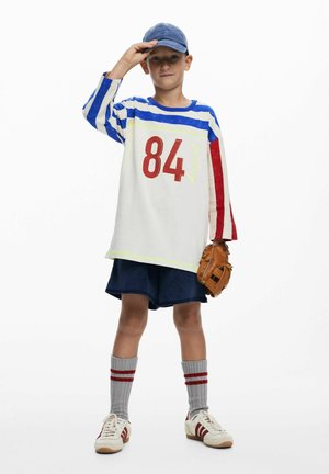 Cămașă cu dungi cu mânecă lungă, cu accente albastre, albe și roșii, având numărul „84” roșu, asortată cu pantaloni scurți bleumarin, șosete gri și mănușă de baseball.