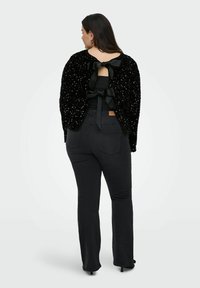 Črn, bleščeč kardigan z odprtim hrbtom in dvema satenastima trakovoma, v kombinaciji s temnimi kavbojkami. Ima sproščen kroj in dolge rokave.