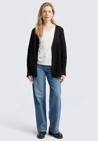 Cardigan côtelé noir avec boutons, associé à un t-shirt blanc à col en V et un jean droit bleu clair. Le mannequin pose les mains sur les hanches.
