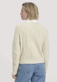 Blonde vrouw die naar links kijkt en een off-white kabeltrui over een wit shirt en blauwe denim jeans draagt, tegen een effen achtergrond.