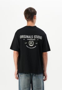 Vyras atsukęs nugarą, vilkintis juodą didelį marškinėlį su baltu užrašu "Originals Studio Copenhagen" ir emblema, derinamas su mėlynomis džinsais.