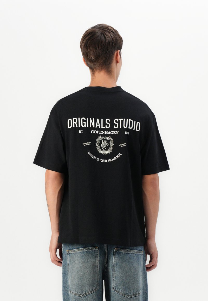 Vyras atsukęs nugarą, vilkintis juodą didelį marškinėlį su baltu užrašu "Originals Studio Copenhagen" ir emblema, derinamas su mėlynomis džinsais.