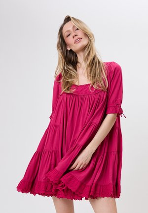 Lyshåret kvinde iført løs, knælængde magenta kjole med puffede ærmer og blonderkant, der poserer mod en ensartet lys baggrund.