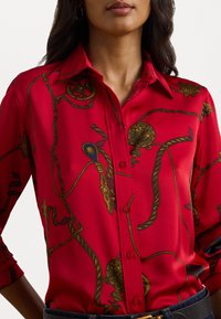 Red silk blouse featuring a detailed pattern of ropes, wheels, and ornamental accents. Classic collar, button-down front, and rolled sleeves.