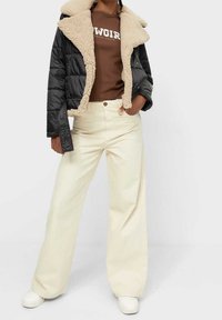 Giacca imbottita nera con collo in ecopelliccia beige, camicia marrone a maniche lunghe con testo bianco, pantaloni larghi color crema e scarpe da ginnastica bianche.