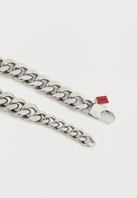 Bracciale a catena in metallo argentato con maglie intrecciate, dotato di una chiusura sicura e di un piccolo tag rosso con la scritta "Hugo". Texture liscia e lucida.
