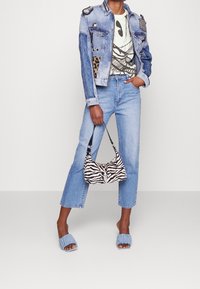Blå denimjakke med dekorative elementer, grafisk t-shirt, lyseblå jeans, zebra-mønstret håndtaske og blå slidesandaler.