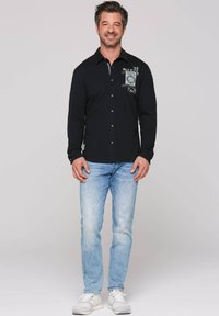 Camisa negra de manga larga con cierre de botones y estampado gráfico en el pecho, combinada con jeans de mezclilla azul claros y zapatillas blancas.
