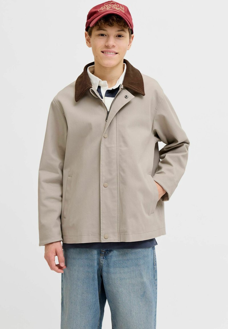 Jeune personne portant une veste beige avec un col marron, une casquette rouge, une chemise blanche à col et un jean bleu clair, debout avec une main dans la poche.