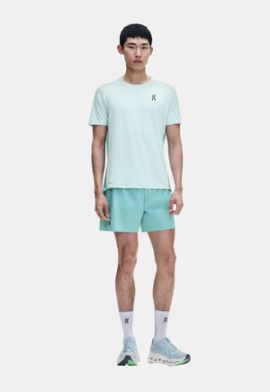 Giovane uomo in piedi indossa una maglietta sportiva azzurro chiaro, pantaloncini abbinati, calzini bianchi e scarpe da corsa blu su uno sfondo semplice.