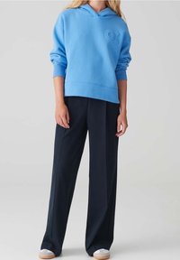 Osoba ve světle modré mikině s embosovaným nápisem "OX", černých širokých kalhotách a bílých teniskách, stojící na jednobarevném pozadí.