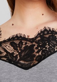 Haut gris avec un col en dentelle noire à motif floral et des détails à franges. Tissu texturé avec une finition lisse en dessous de la dentelle.