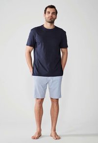 T-shirt en coton bleu marine associé à un short bleu clair. Les deux présentent une coupe décontractée et une texture douce. Le modèle est pieds nus avec les mains dans les poches.
