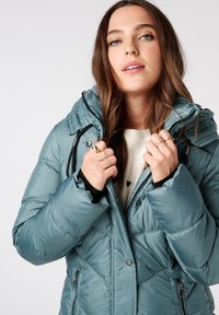 Dikke teal jas met chevronstiksel, verstelbare capuchon, ritssluiting aan de voorkant en drukknoopsluiting, gedragen door een persoon in een licht sweater.