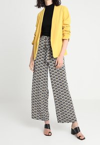 Blazer jaune, top noir ajusté, pantalon large à motifs bleu et or, et sandales à talons noires. Tissu lisse avec une coupe décontractée.