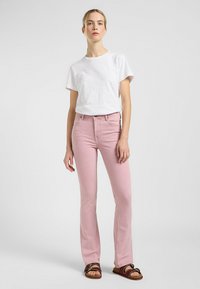 Roze flared jeans met een gladde textuur en vijf-pocket ontwerp, gecombineerd met een wit katoenen t-shirt. Model draagt bruine sandalen.