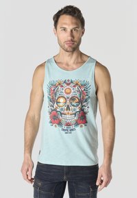 Hombre con camiseta sin mangas azul claro con calavera de azúcar colorida, flores tropicales, aves y texto "Tropic Vibes", combinado con vaqueros oscuros.