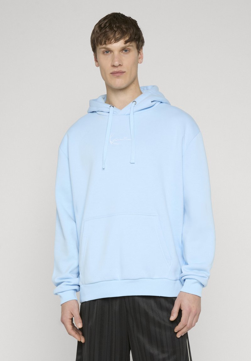 Jeune homme portant un sweat à capuche bleu clair avec poche avant et pantalon plissé noir, debout devant un fond clair uni.