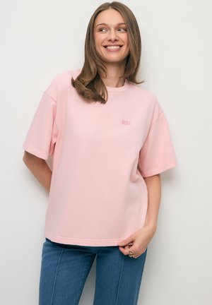 Vrouw draagt een lichtroze oversized T-shirt en blauwe jeans, glimlacht en kijkt naar de zijkant tegen een effen lichte achtergrond.