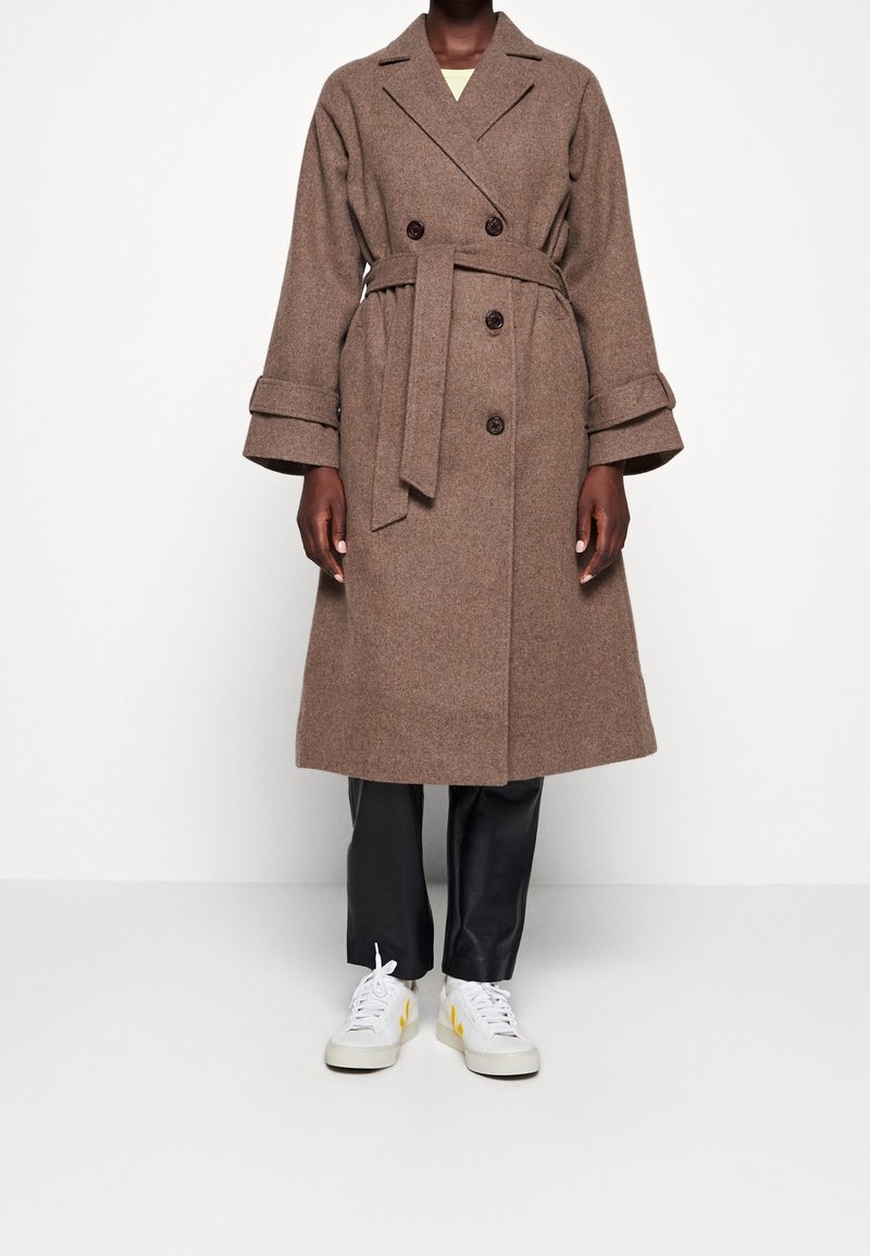 Brun uldblanding frakke med dobbeltrækknaplukning, brede ærmer og bælte i taljen. Kombineret med sorte bukser og hvide sneakers.