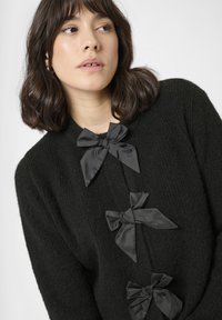 Sort strikket sweater med tre store satinstof-sløjfer fordelt langs fronten, med et tekstureret design og en høj halsudskæring.