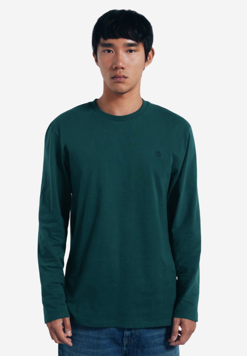 Langärmliges grünes Shirt aus weichem Stoff mit rundem Halsausschnitt und subtiler Logodarstellung auf der Brust. Kombiniert mit Jeans.