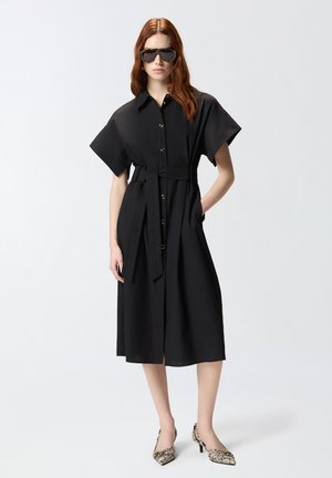 Donna con lunghi capelli rossi che indossa un vestito chemisier midi nero con cintura, occhiali da sole oversize e tacchi alti in pelle di serpente, in piedi su sfondo bianco.