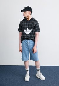 Juodos pltadėlės trumpomis rankovėmis marškiniai su balta Adidas emblema, šviesiai mėlyni džinsiniai šortai, juoda beisbolo kepurė, balti sportbačiai ir dryžuotos kojinės.