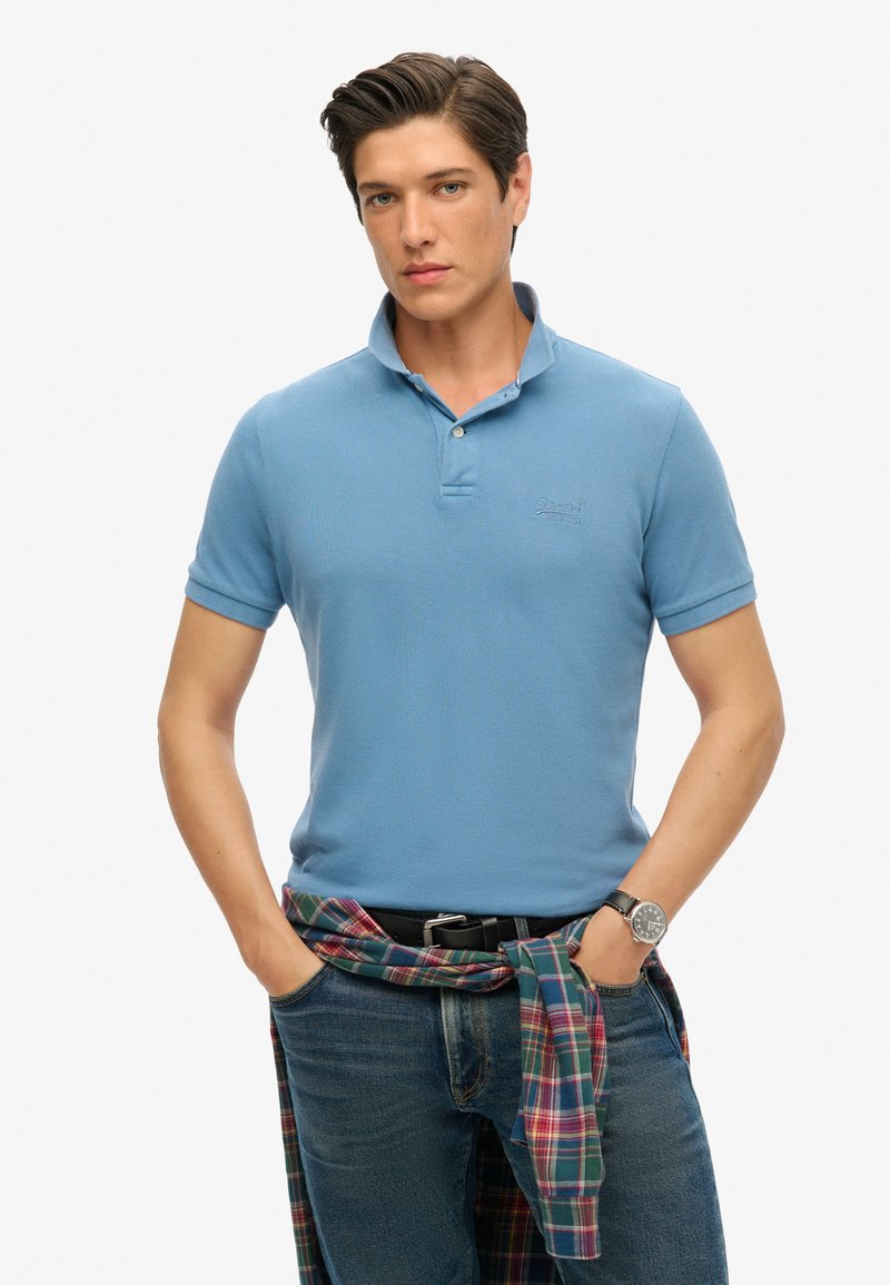 Polo shirt azzurro chiaro a maniche corte con un motivo testurizzato, patta con due bottoni e logo ricamato; abbinato a jeans scuri e una camicia a quadri legata in vita.