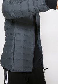 Persona che indossa una giacca imbottita grigio scuro su abbigliamento nero con pantaloni a righe bianche, braccio alzato, vista laterale parziale su sfondo semplice.
