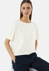 Witte T-shirt met korte mouwen van zacht katoen. Beschikt over een ronde halslijn en een klein borstzakje. Gecombineerd met donkerblauwe broek.