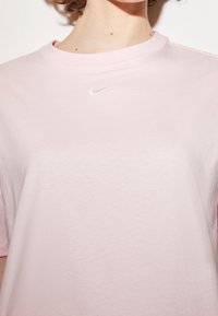 Světle růžové bavlněné tričko s kulatým výstřihem; na levé straně hrudi se nachází malé bílé logo Nike; hladká textura a uvolněný střih.