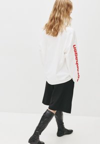 Hvid langærmet top med rød "Copenhagen" tekst på ærmet, parret med sorte knælange shorts og glinsende sorte støvler.