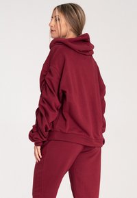 Bordowa bluza z kapturem o luźnym kroju, z marszczonymi rękawami, sznurkiem do regulacji kaptura oraz miękką teksturą, w komplecie ze spodniami dresowymi w tym samym kolorze.