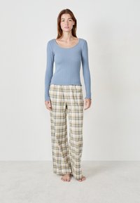 Femme debout pieds nus, portant un haut à manches longues côtelé bleu clair et un pantalon large à carreaux beiges, sur fond uni.