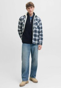 Camisa de franela a cuadros con un cuello de forro polar blanco, superpuesta sobre un suéter de punto azul marino, combinada con jeans azul claro y calzado beige.