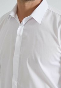 Chemise blanche avec une texture lisse et sans plis, dotée d'un col pointu et d'une patte de boutonnage discrète dans un style classique ajusté.